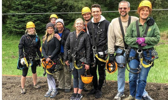 Group of eight adults wearing yellow helmets and safety harnesses posing outdoors, promoting zip-lining or adventure activities available at the Cape May or Lewes destination.