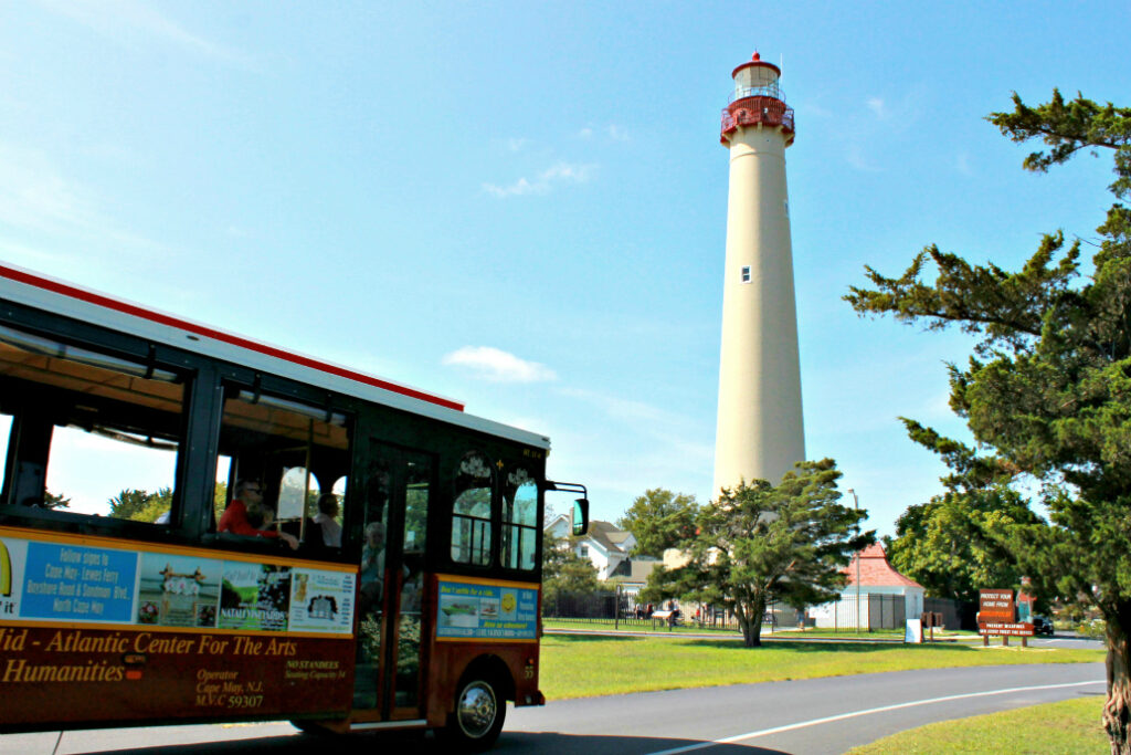 Photo of a local trolley/shuttle approaching a tall lighthouse- creme colored with a red roof/deck near the top- promoting Cape May, NJ museums