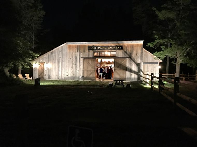 Exterior of the Cold Spring Brewery barn at night, featuring a brightly lit open entryway revealing patrons inside and a wooden split-rail fence in the foreground, promoting local breweries near the ferry.