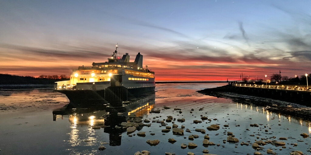 Cape May-Lewes Ferry illuminated at twilight, departing the terminal over water with reflecting ice floes and a dramatic red and orange sunset in the background.