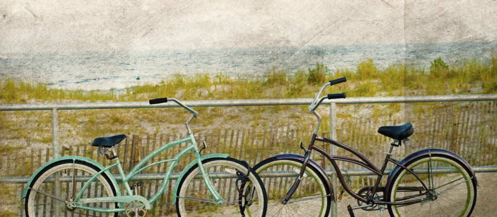 Two retro cruiser bicycles, one mint green and one brown, parked by a metal fence with beach dunes and the ocean visible in the background, promoting cycling near the ferry terminals.