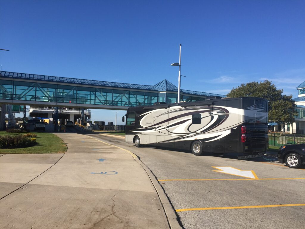 Large black and white Class A motorhome RV preparing to board the Cape May-Lewes Ferry at the terminal entrance, showcasing the glass pedestrian walkway above and promoting ferry travel for large vehicles.