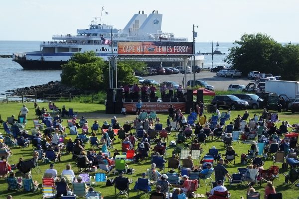 Concert-goers sit on their lawn chairs on the "green" next to the Cape May Ferry terminal in front of the stage, with the Ferry sailing in the background